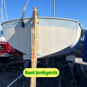 Front view of a white sailboat hull on supports in a boat yard, with masts in the background and the ‘Boat Junkyards’ logo at the bottom edge.
