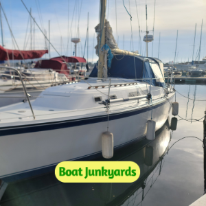 Sailboat moored at a marina with a blue canopy on the cockpit and a green Boat Junkyards sticker on the hull.
