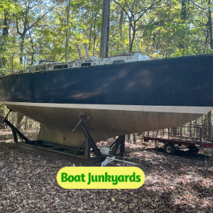 Blue boat on a trailer in a wooded yard with fallen leaves and a Boat Junkyards logo in the foreground.