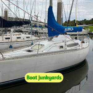 White sailboat docked in a marina with blue sail cover, visible masts, and a Boat Junkyards logo on the hull line.
